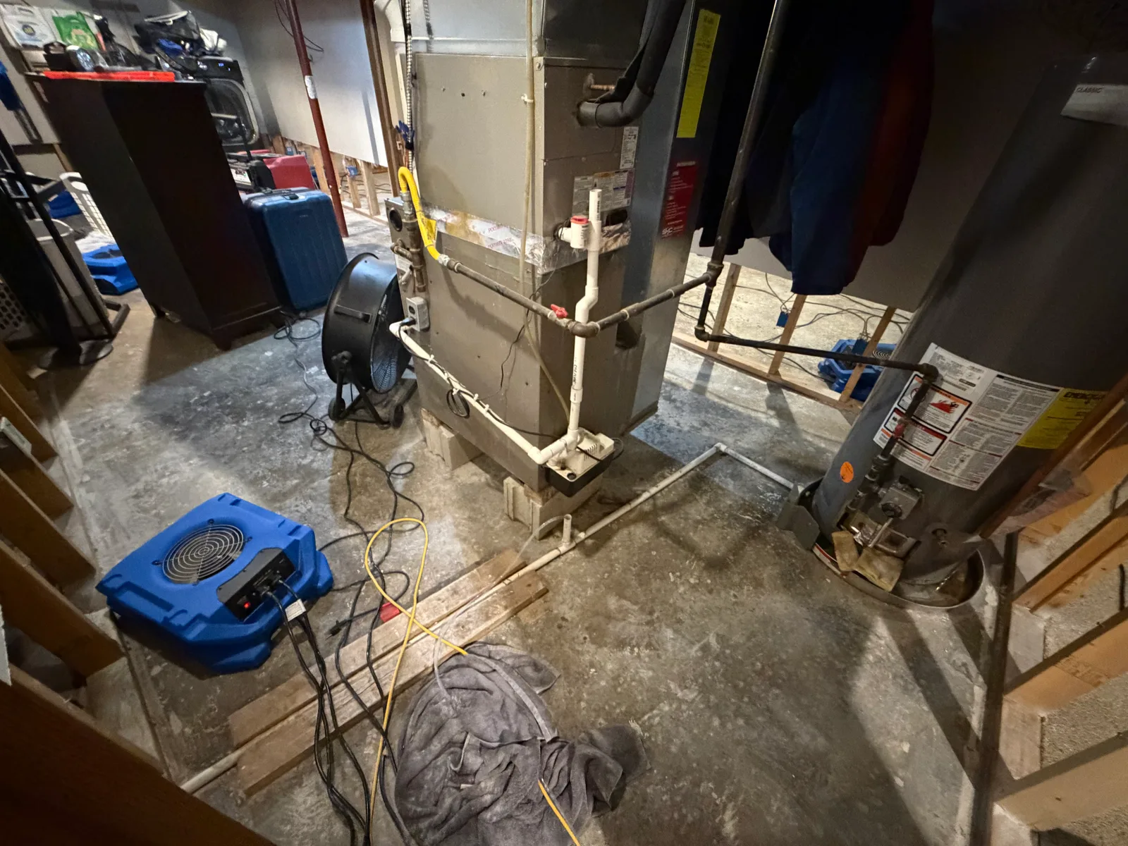 A basement with a blue air mover and a black drying fan positioned on a damp concrete floor near a furnace and water heater, with exposed subfloor and scattered cords.