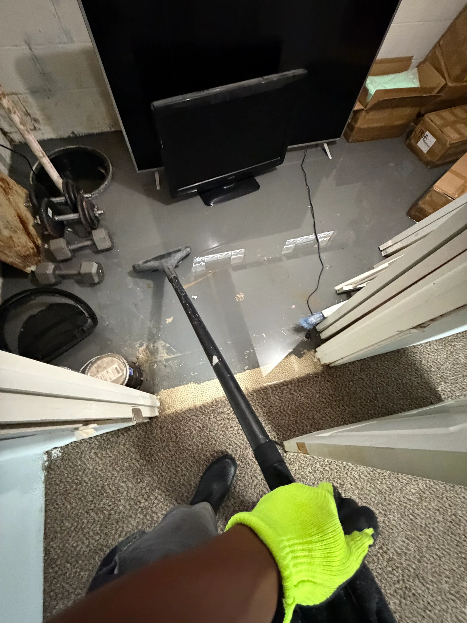 A gloved hand holds a long-handled squeegee over a wet basement floor with standing water near a television, dumbbells, boxes, and stacked trim.