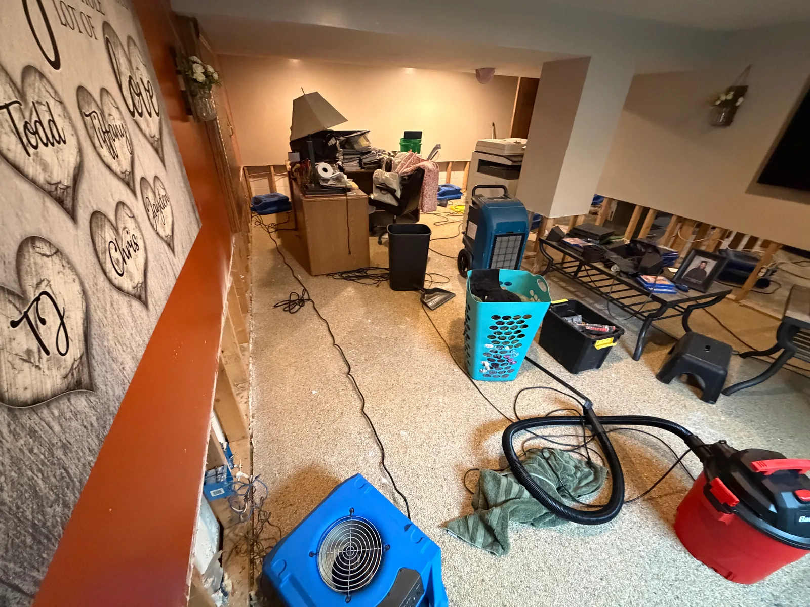 A basement with water damage restoration equipment including a blue air mover, dehumidifier, and red wet/dry vacuum, with removed lower drywall exposing wall studs and cluttered furniture.
